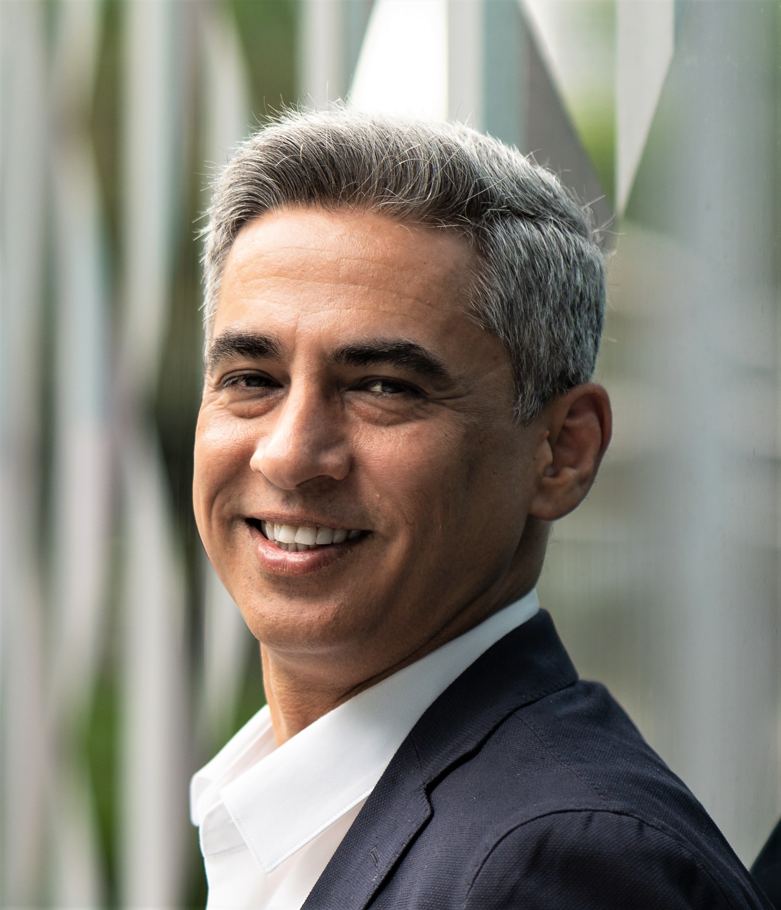 A man with short gray hair and a slight smile is wearing a dark suit jacket and white collared shirt, standing outdoors with a blurry geometric background.
