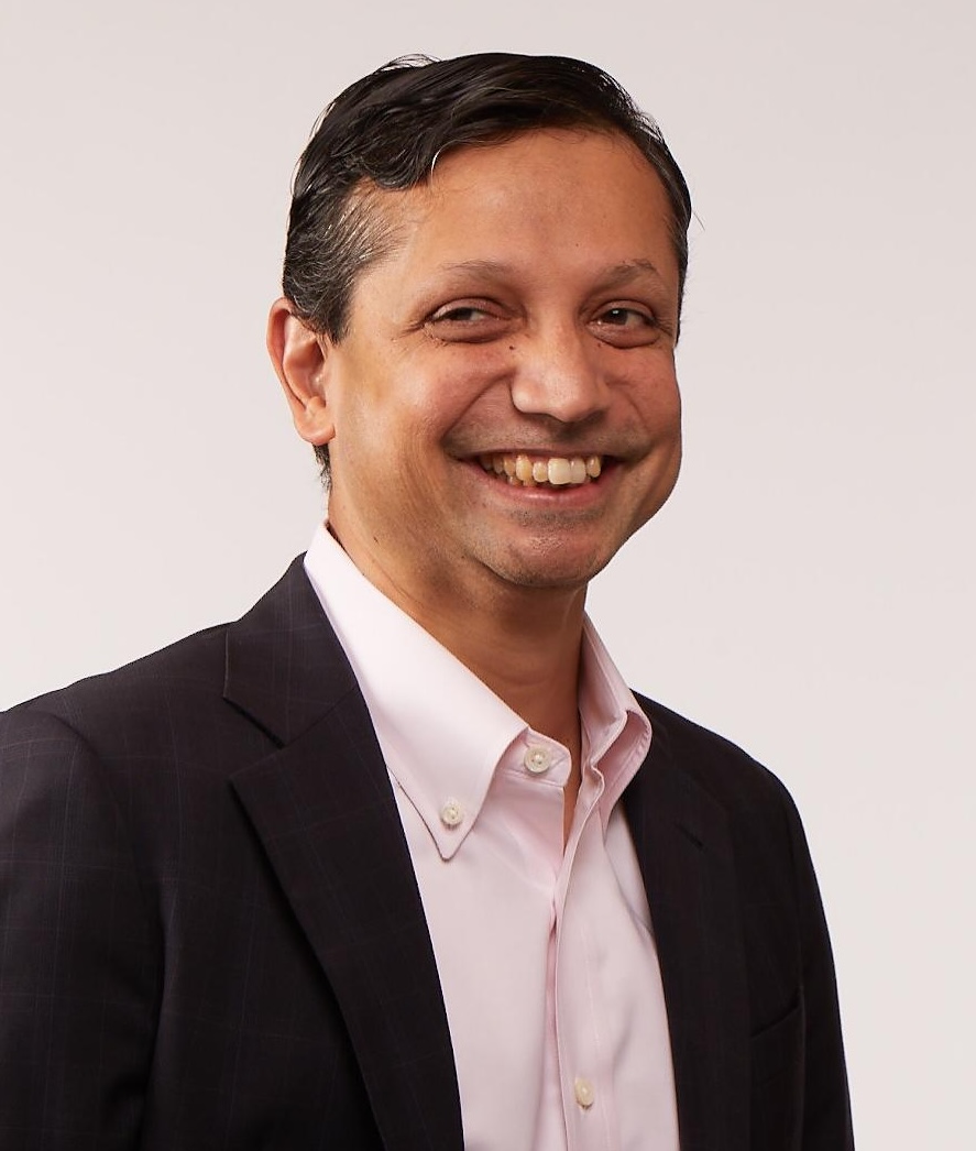 A man in a dark suit and light pink shirt smiles while looking slightly to the left against a plain background.