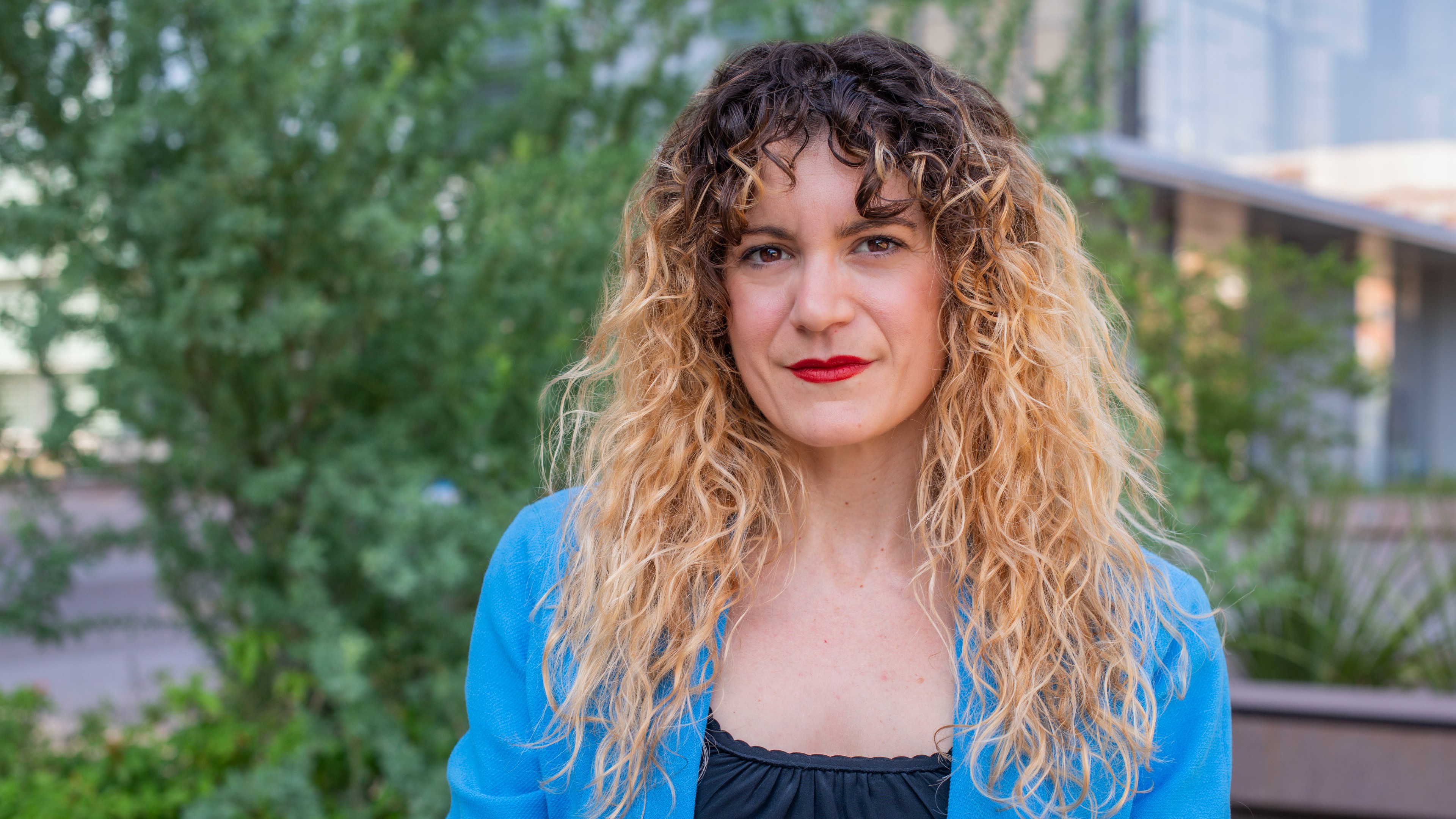 A person with curly hair and a blue blazer poses outdoors in front of greenery.
