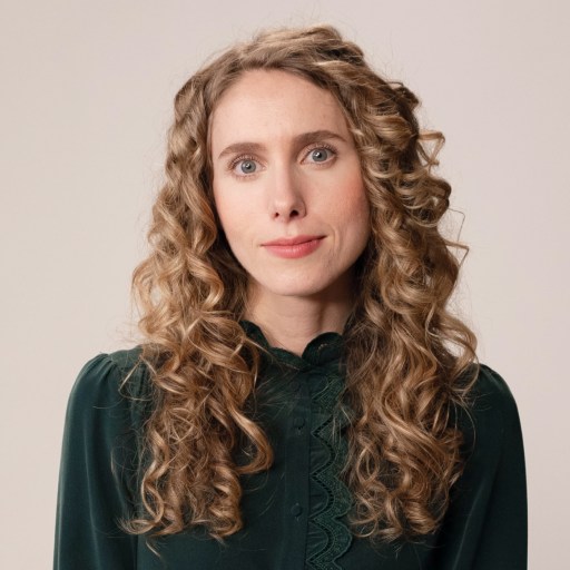 A person with long, curly blonde hair wearing a dark green blouse stands against a plain background.