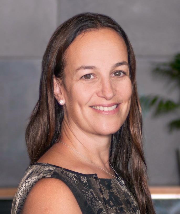 A woman with long brown hair, wearing a patterned top, smiles at the camera with a neutral, indoor background.