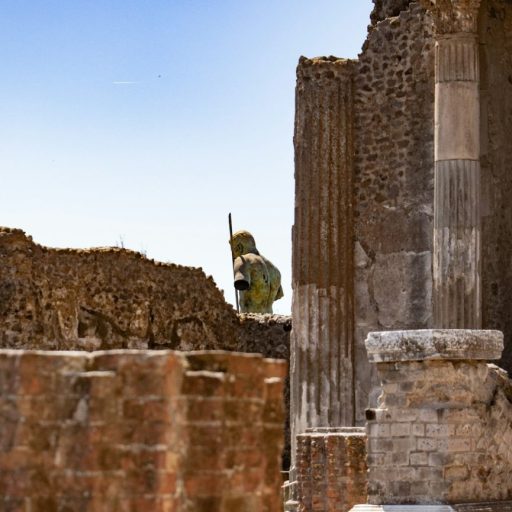 Ancient ruins with stone columns and damaged walls, featuring a statue partially visible in the background. Clear blue sky above.