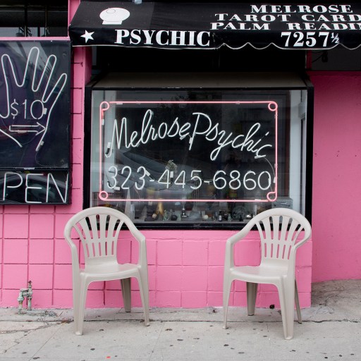 A storefront with signs for psychic telepathy readings and a phone number. Two white plastic chairs are placed in front. The storefront is pink with a black awning and a large hand sign advertising $10 readings.