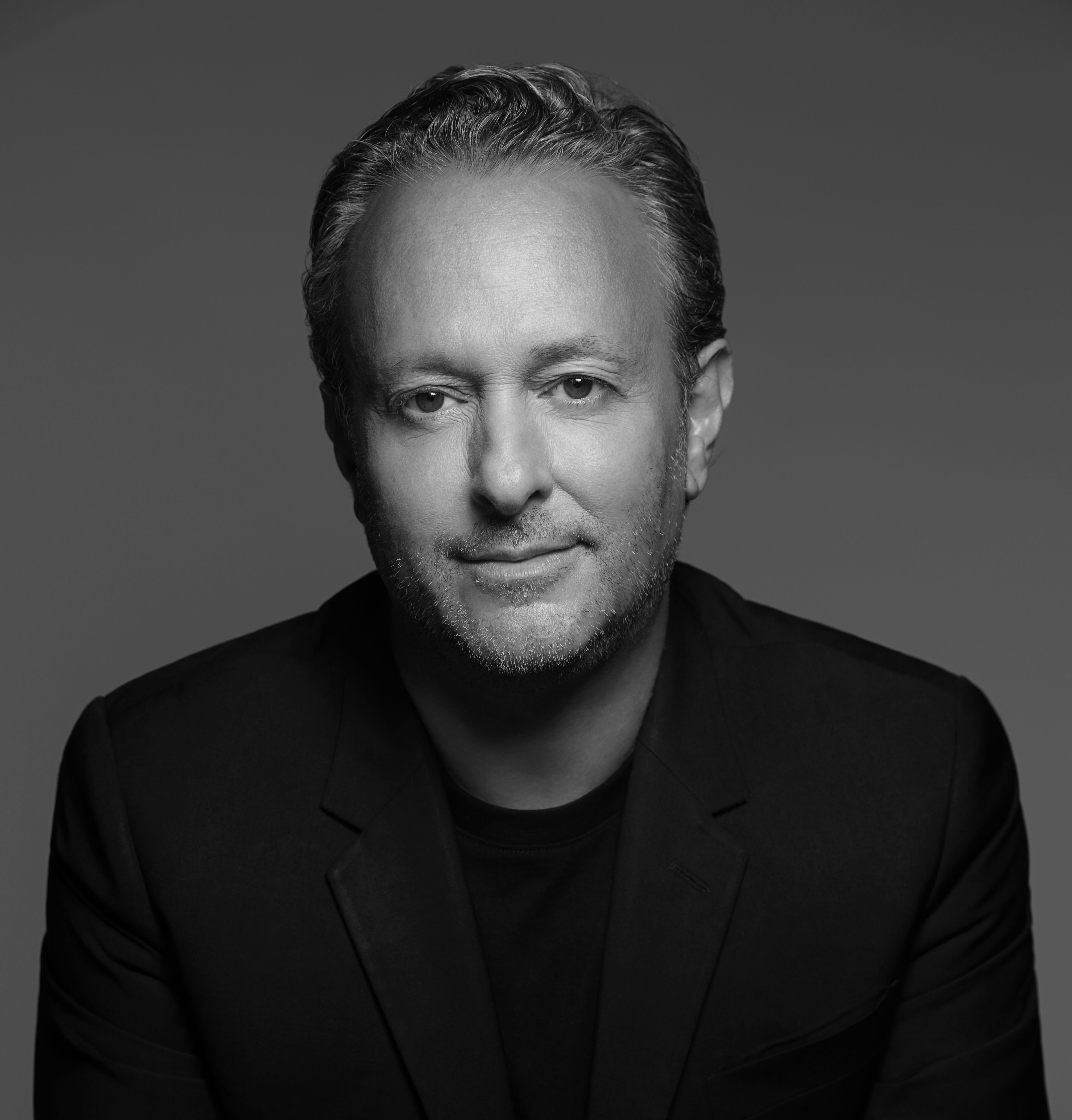 Black and white portrait of a middle-aged person with short, curly hair wearing a dark blazer, smiling slightly.