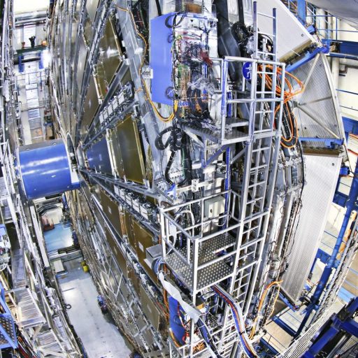 A large, intricate machine with metallic components and blue scaffolding in a laboratory setting. Numerous cables and pipes are connected to the central structure.