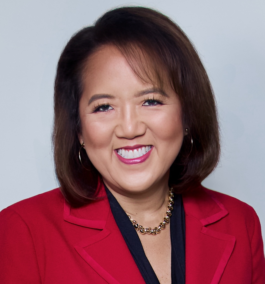 A person with shoulder-length brown hair, wearing a red blazer and necklace, smiles against a plain background.
