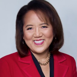 A person with shoulder-length brown hair, wearing a red blazer and necklace, smiles against a plain background.