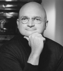 A bald man wearing round glasses and a dark sweater poses with his hand on his chin. The background is blurred.