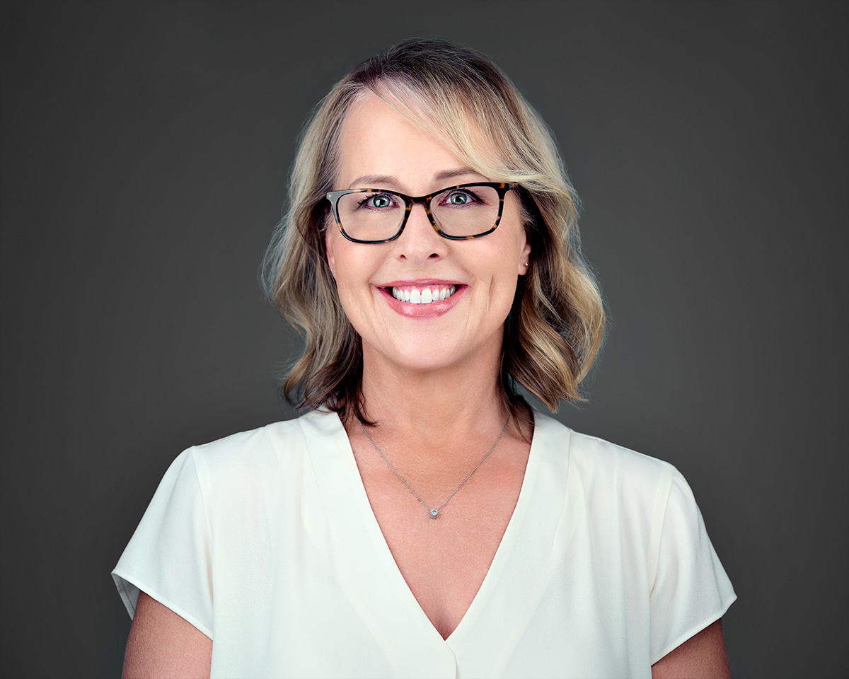 A woman with blonde hair and glasses is smiling. She is wearing a white blouse with a small pendant necklace. The background is plain and dark.