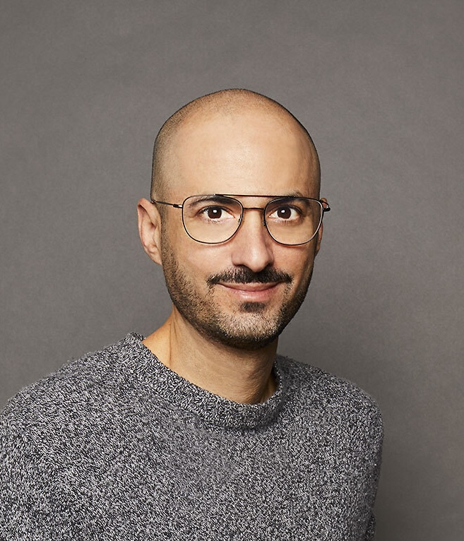 A man with glasses, a beard, and a shaved head, wearing a gray sweater, smiles against a plain gray background.