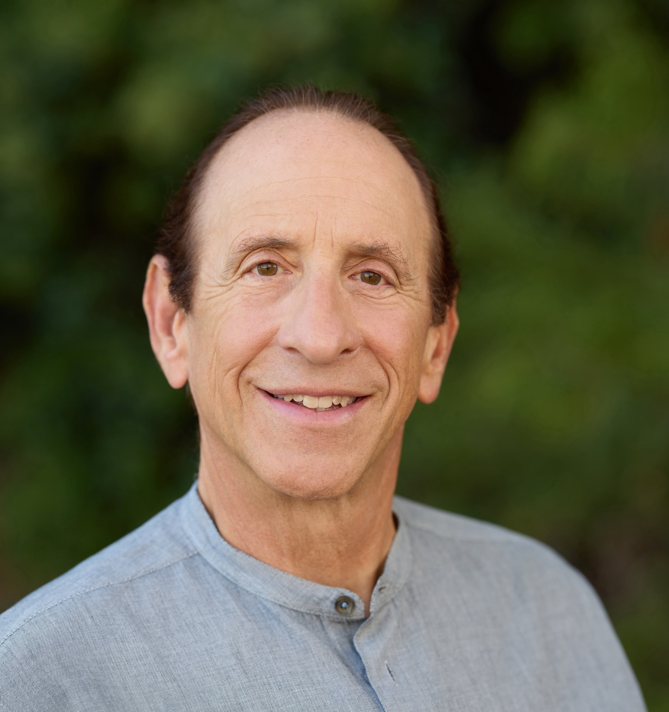 A man with short, dark hair smiles at the camera while wearing a light gray shirt in an outdoor setting with greenery in the background.