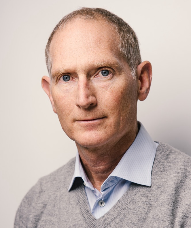 A middle-aged person with short gray hair, wearing a light blue shirt and gray sweater, looking directly at the camera.