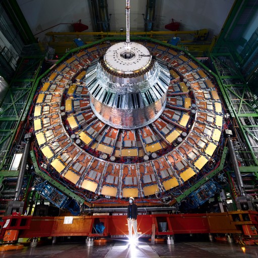A person stands in front of a large, circular particle detector in a brightly lit, high-tech facility.