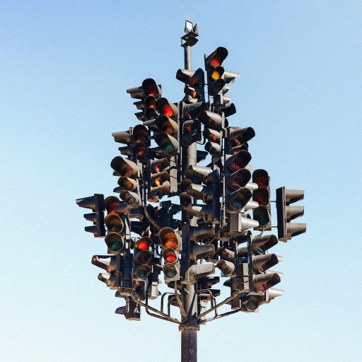 A pole with numerous traffic lights mounted in different directions creates an illusion of control against a clear sky background.