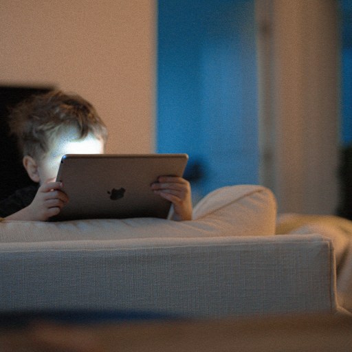 Young child sitting on a couch, illuminated by the screen of a tablet they are holding in a dimly lit room.