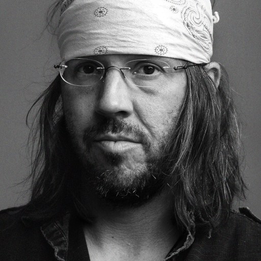 A man with long hair, a beard, glasses, and a bandana on his head, reminiscent of David Foster Wallace, poses for a portrait against a plain background.