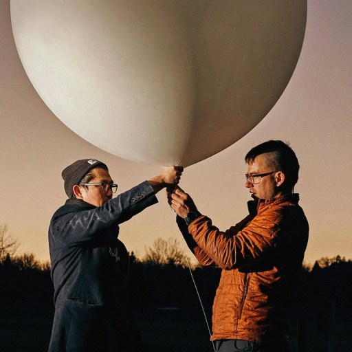 Two individuals are holding and inspecting a large white balloon outdoors, with a silhouette of trees visible in the background at dusk.