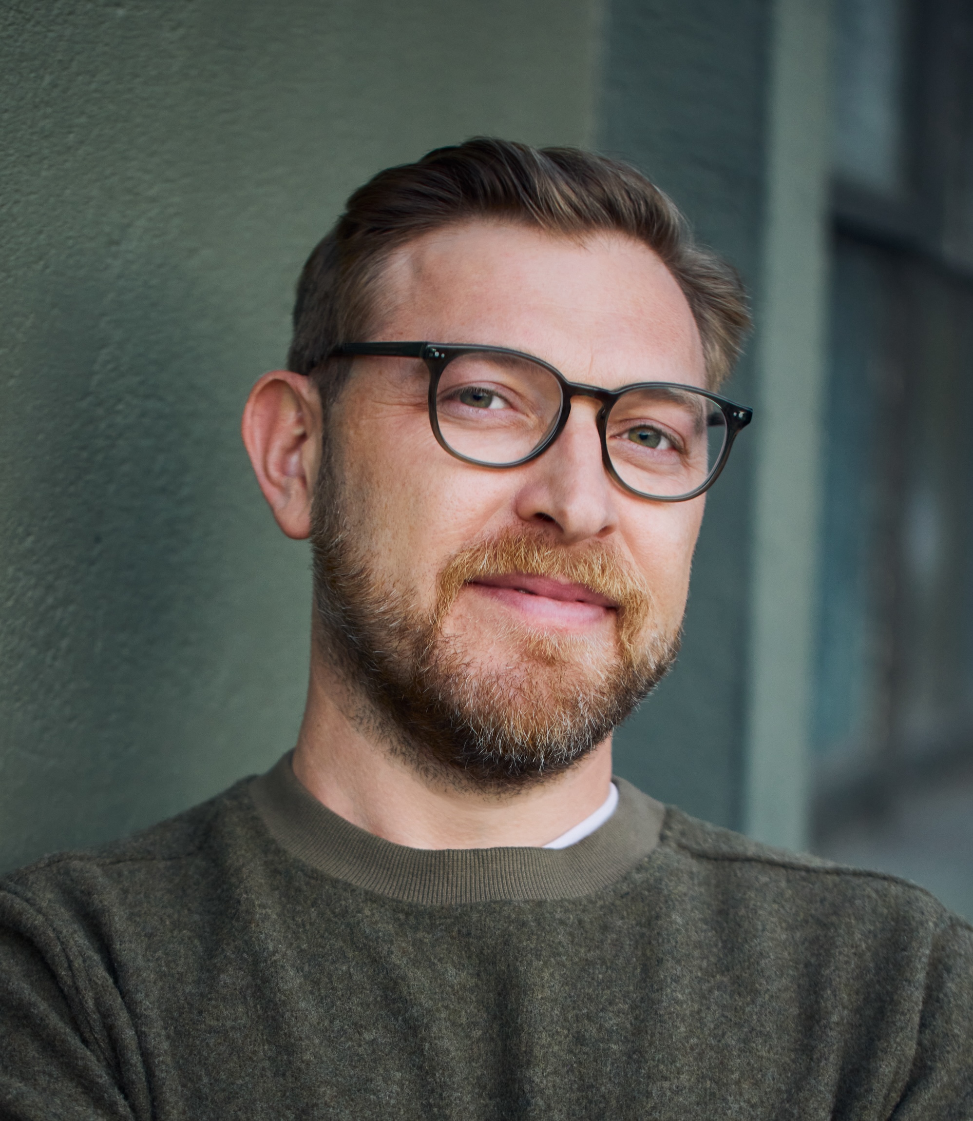 Man with glasses and a beard, wearing an olive sweater, leans against a textured wall and smiles slightly.