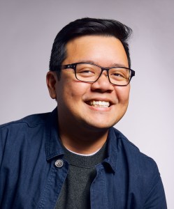 A person wearing glasses and a dark sweater smiles against a light gray background.