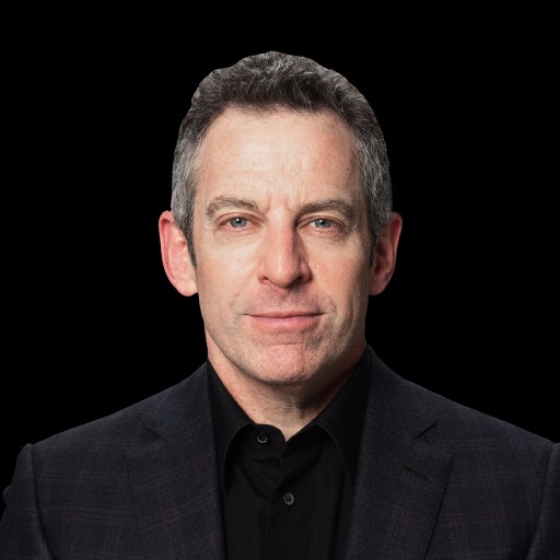 A man with short, grayish hair, wearing a dark blazer and black shirt, stares directly at the camera against a black background.