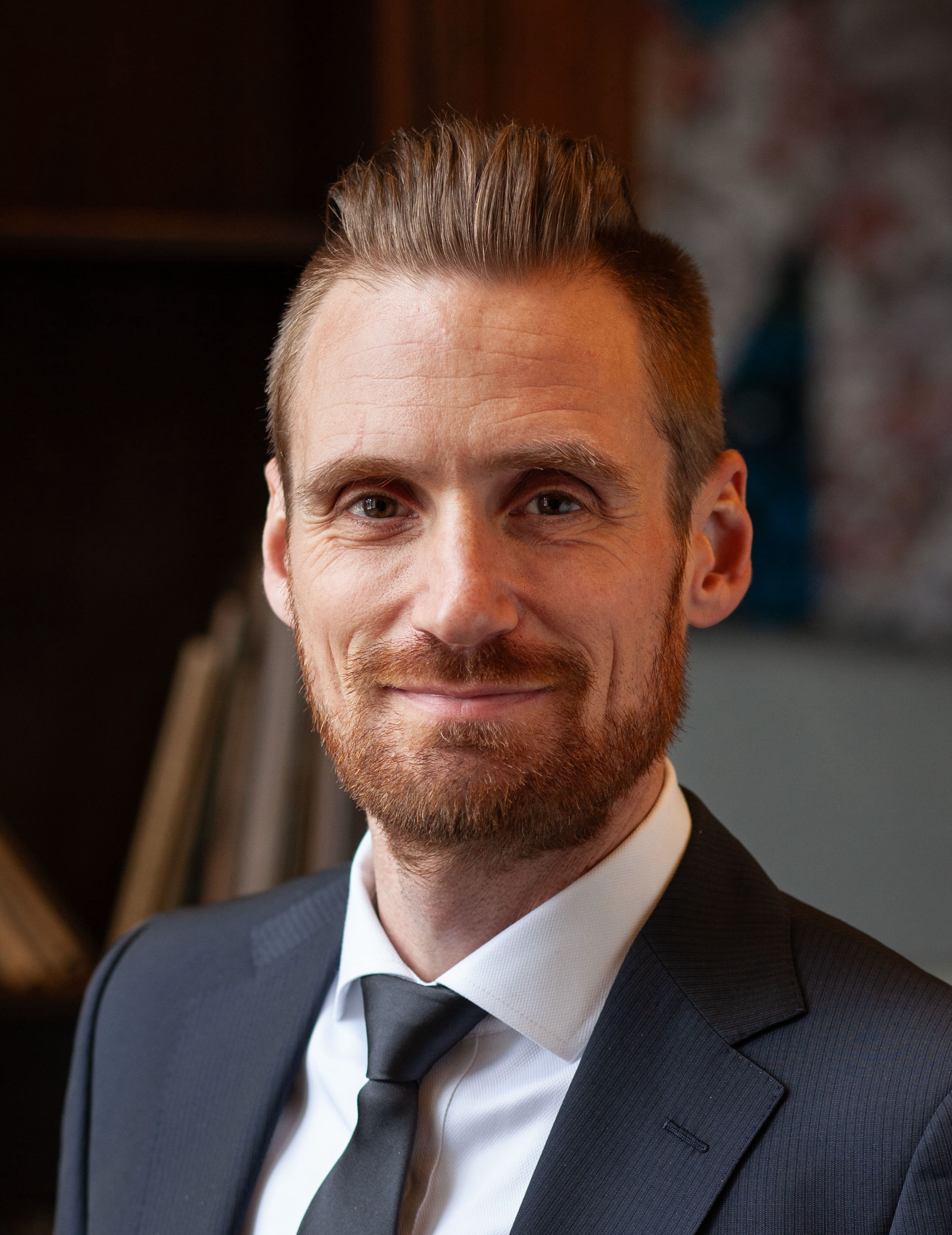 A man with short hair and a beard wears a suit and tie, smiling slightly, in an indoor setting.
