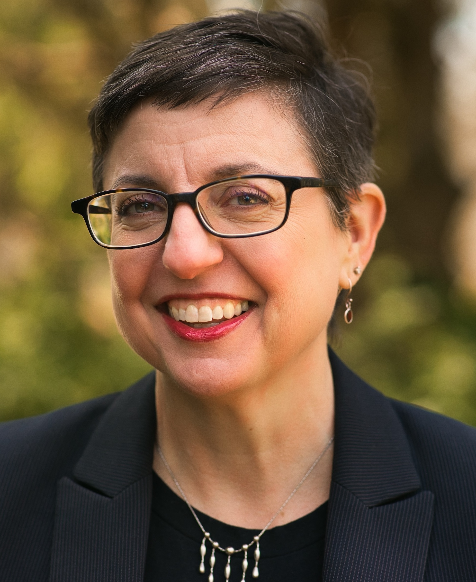 A person with short hair and glasses smiles, wearing a black blazer and earrings, standing outdoors.