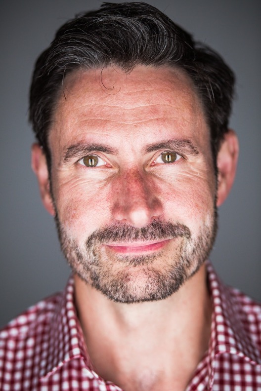A man with a beard and mustache, wearing a red and white checkered shirt, looks directly at the camera against a gray background.