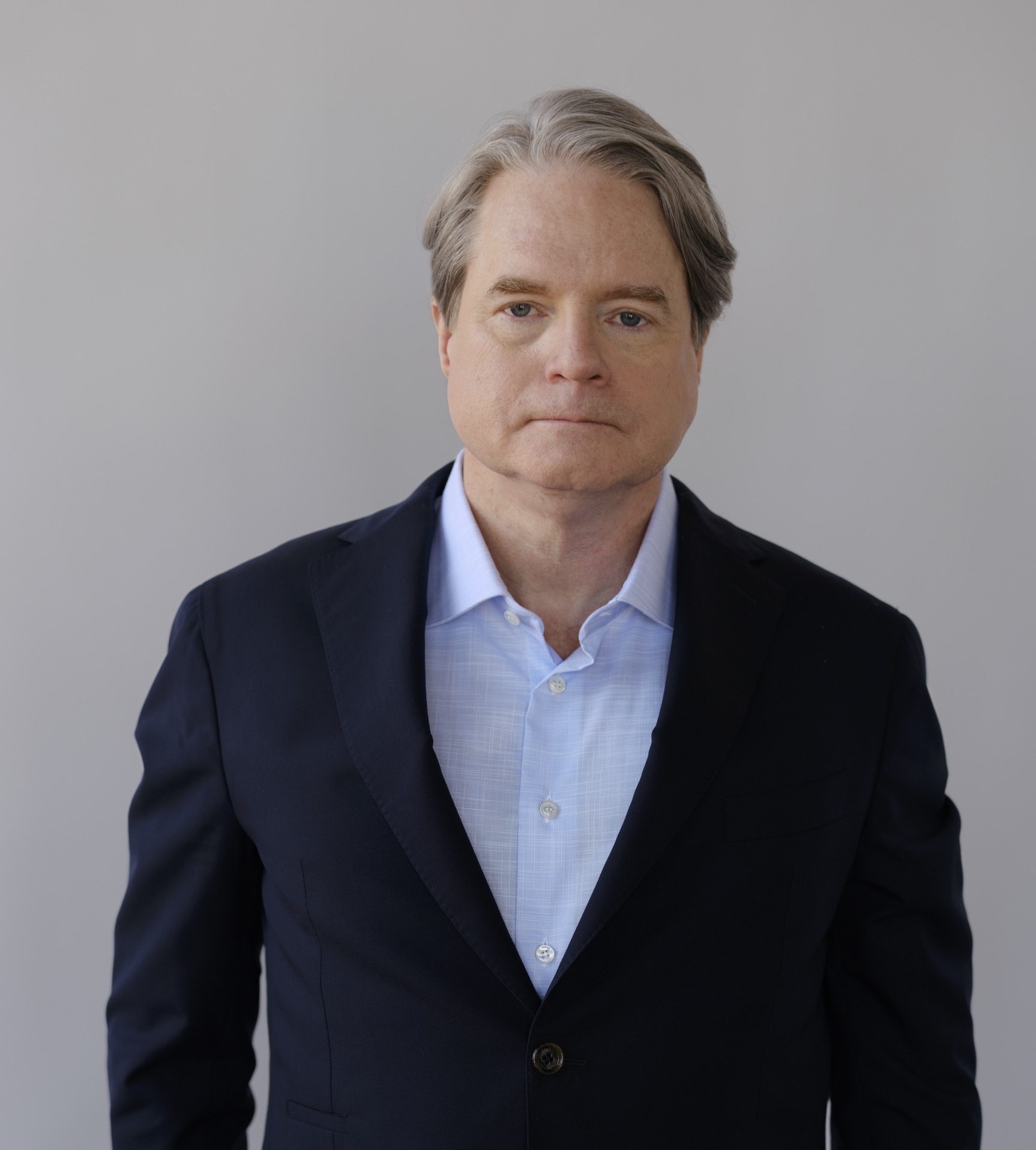 A man in a dark suit and light blue shirt stands against a plain background.