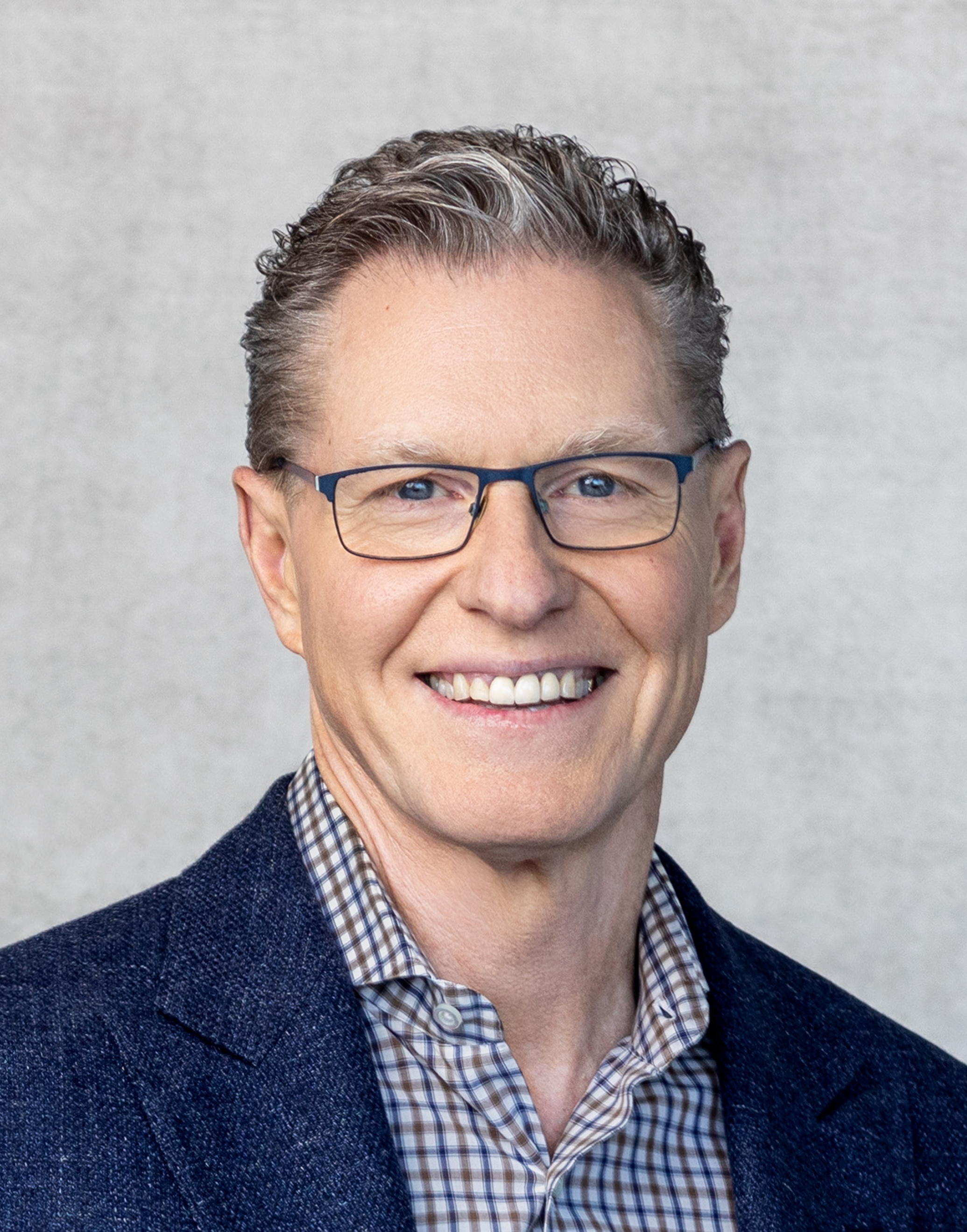 Man with glasses smiling, wearing a blazer over a checkered shirt against a plain background.