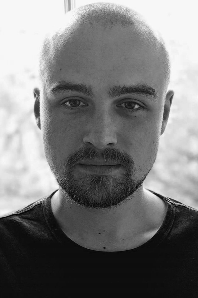 A person with a shaved head and beard looks directly at the camera, wearing a dark shirt. The background is softly blurred. Black and white image.