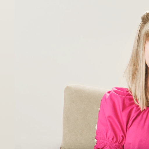 A woman with blonde hair smiles while sitting on a beige chair. She wears a bright pink top and the background is a plain light color.