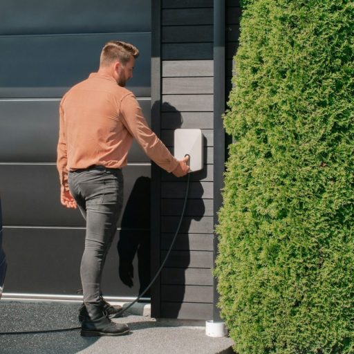 A man connects an electric car to a charging station mounted on a house wall next to a large shrub.