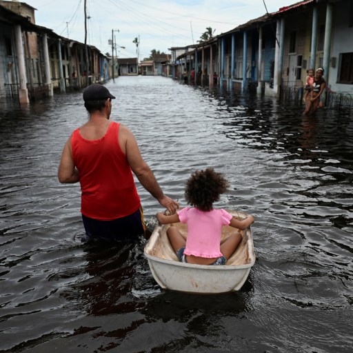 Amidst the chaos of natural disasters, a man pulls a child in a small boat through a flooded street lined with houses.