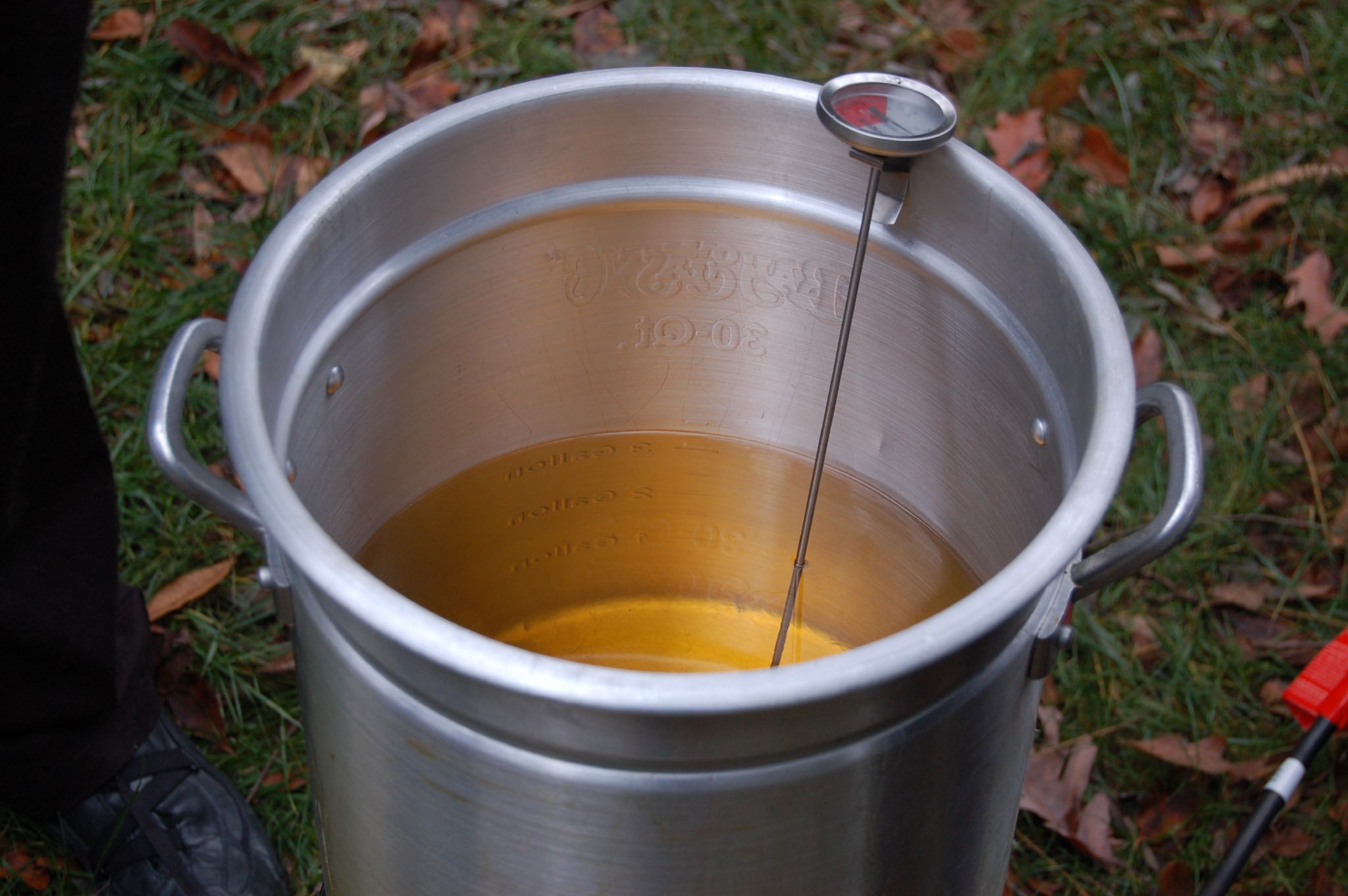 A metal pot filled with oil, along with a thermometer, sits on the grass, ready to safely deep fry a frozen turkey.