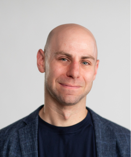 A person with a shaved head, wearing a dark blazer and black shirt, smiles slightly against a neutral background.