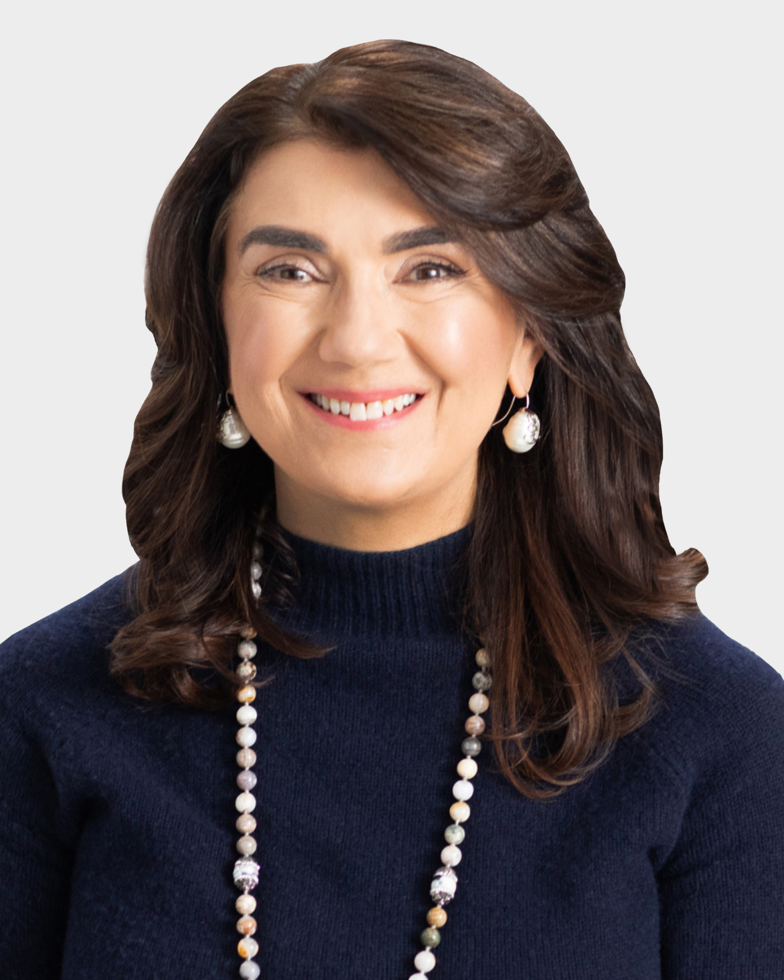 A woman with long dark hair and pearl earrings smiles while wearing a dark sweater and a beaded necklace against a plain background.