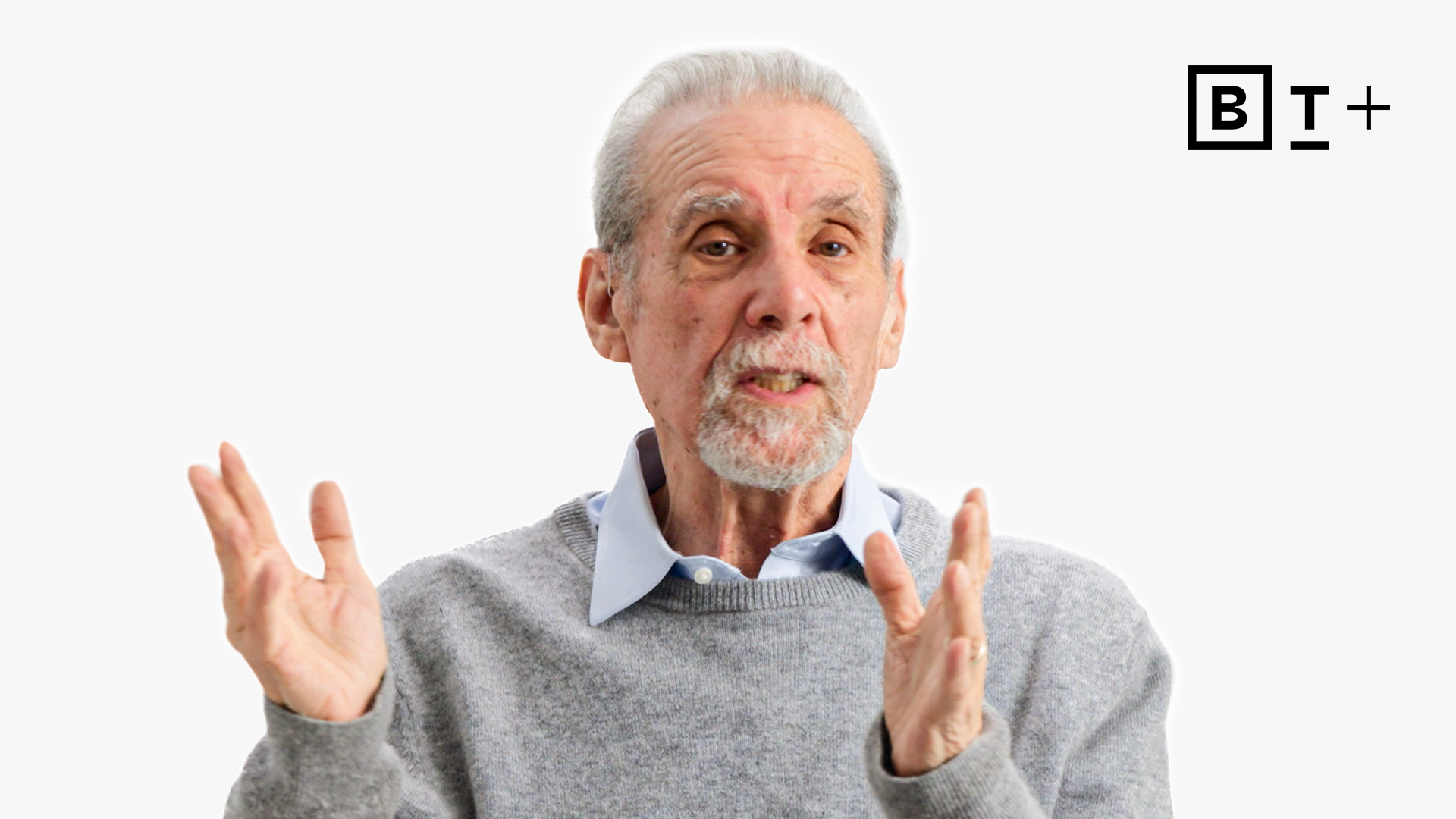 An elderly man gestures with his hands while speaking, wearing a gray sweater and blue shirt. The background is plain white with a logo in the top right corner.