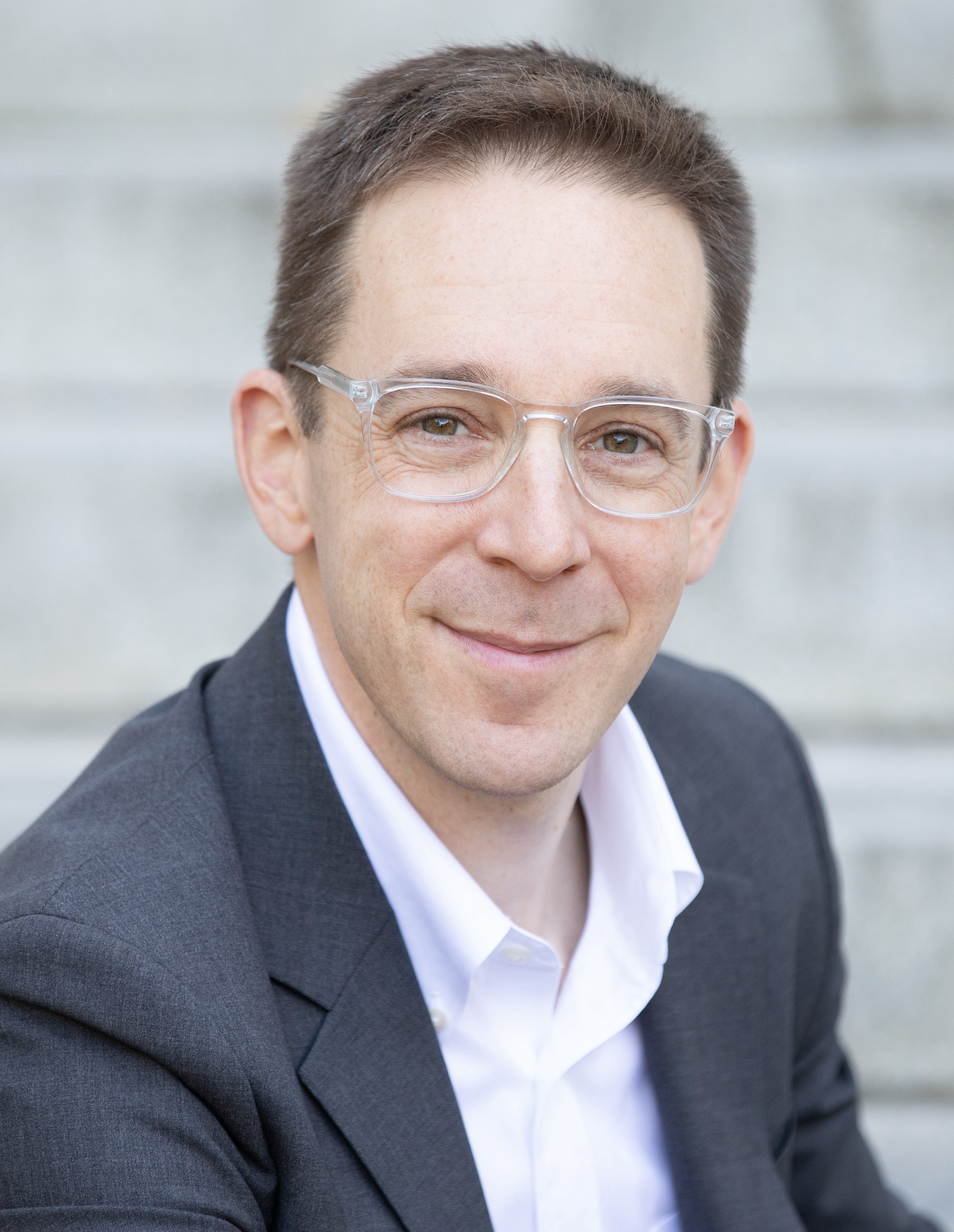 A person in a suit and glasses smiling, sitting on steps.