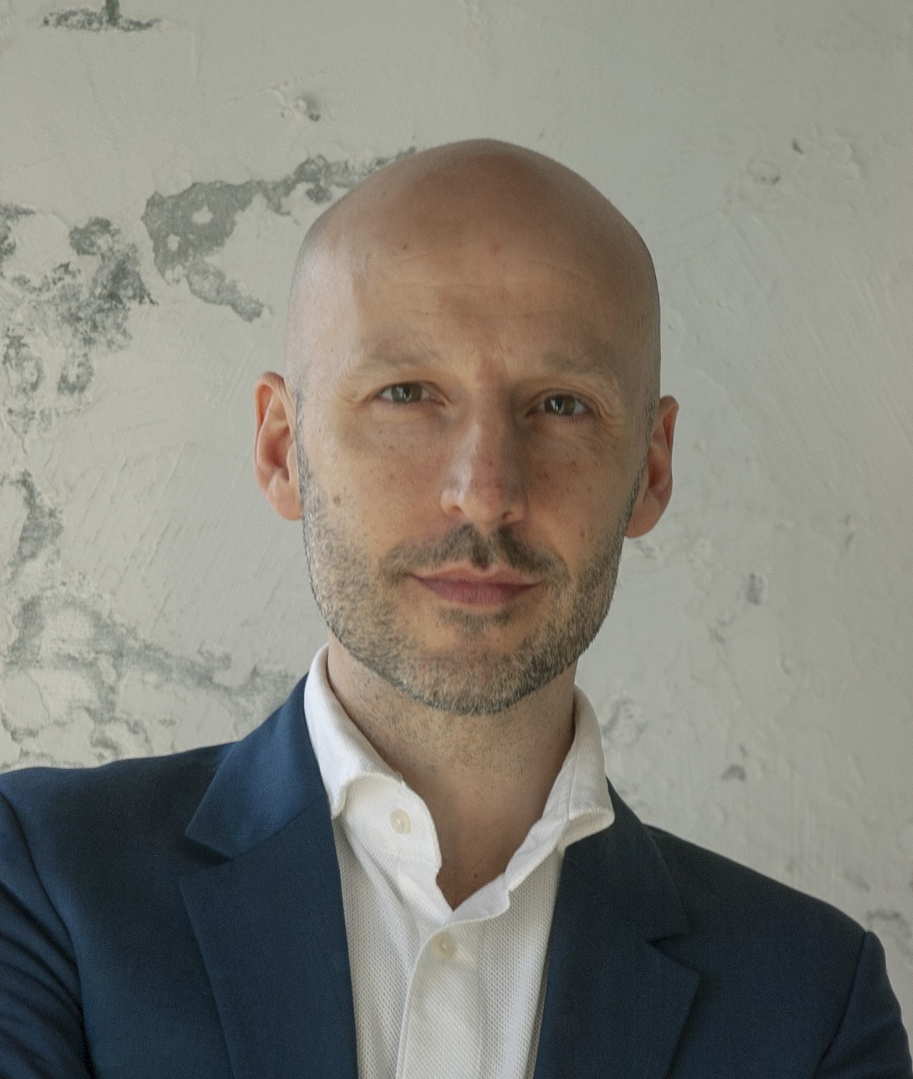 A bald man with a beard wearing a suit and white shirt stands in front of a textured wall.