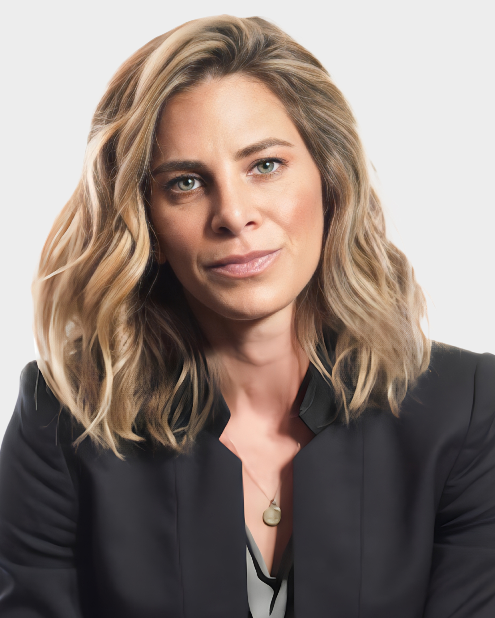 A person with wavy, shoulder-length hair is wearing a black blazer and a pendant necklace, facing forward against a plain background.