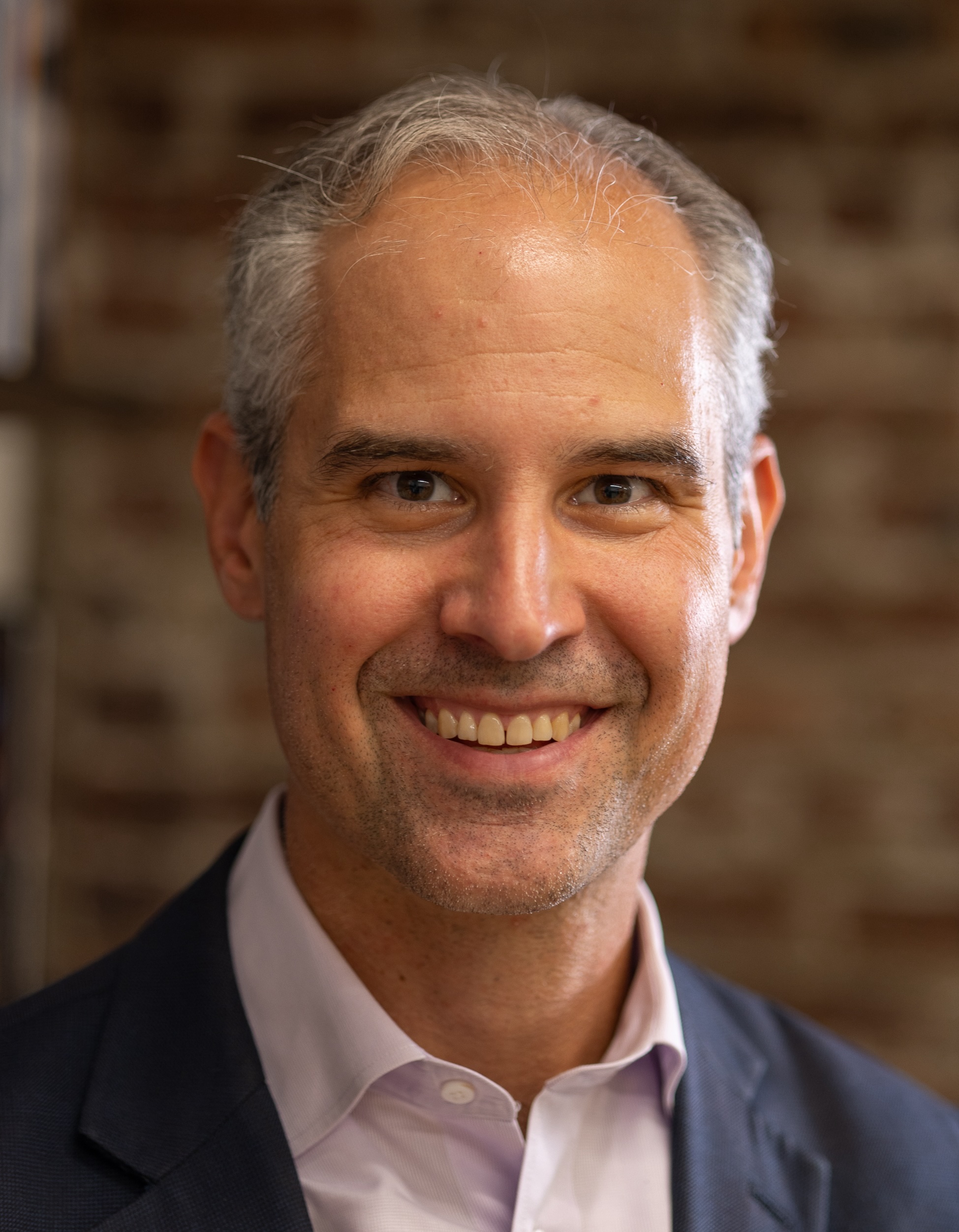 A person with short gray hair and a suit smiling in front of a blurred brick wall.