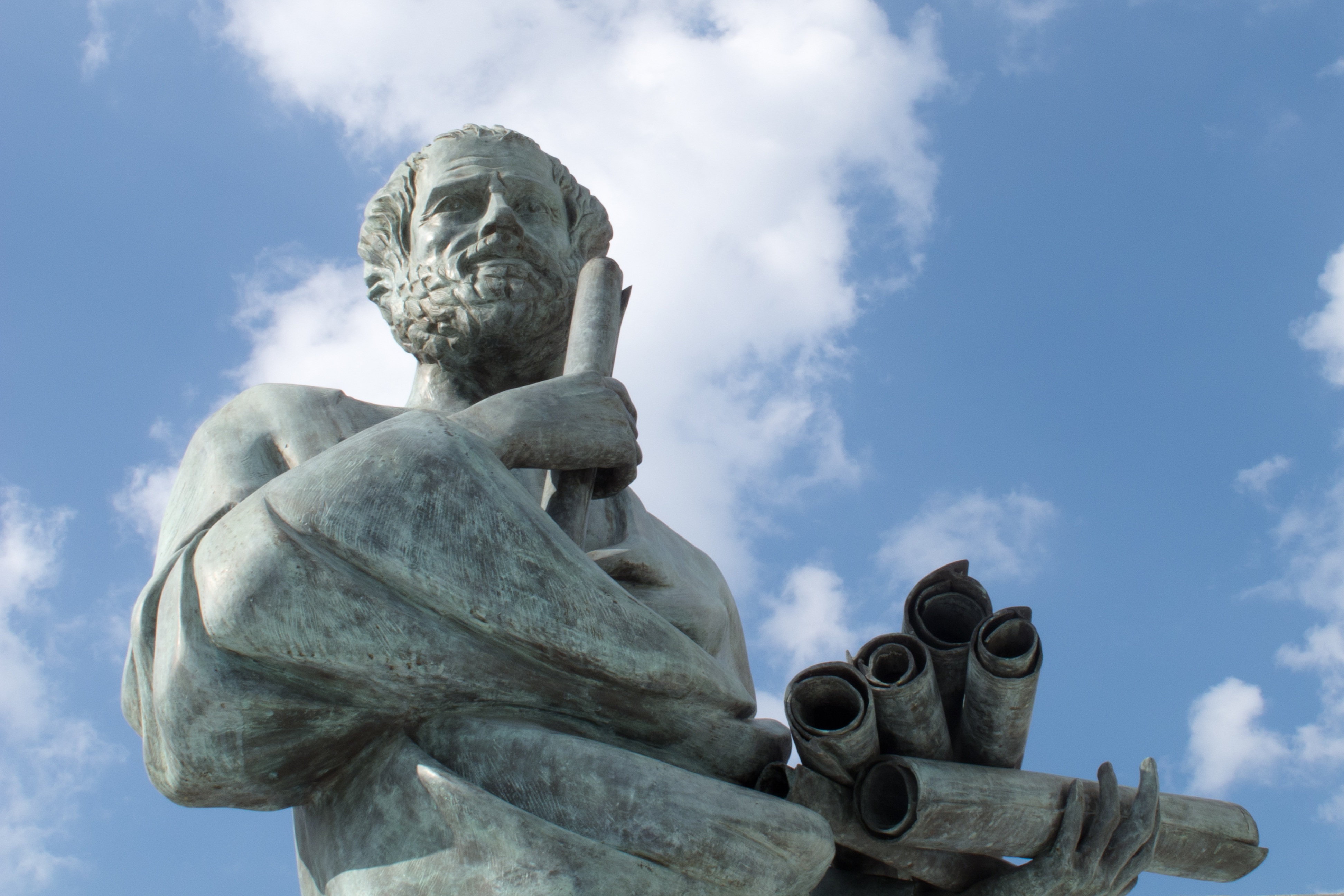 A bronze statue of a bearded man holding scrolls against a backdrop of a blue sky with clouds.
