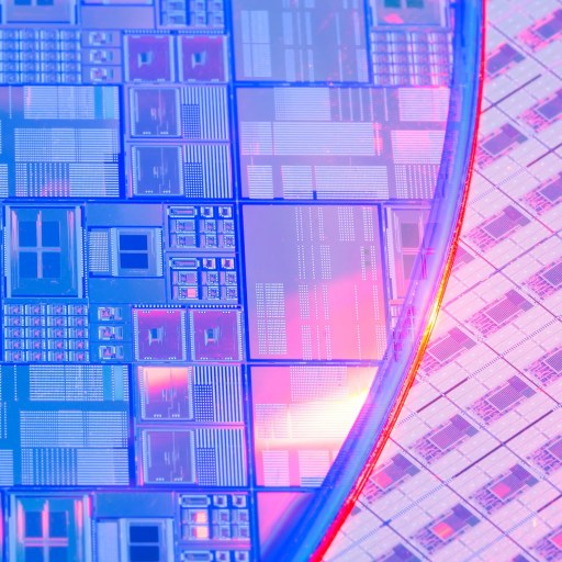 Close-up of a semiconductor wafer with intricate microchip patterns and a reflective surface, illuminated by pink and blue lights.