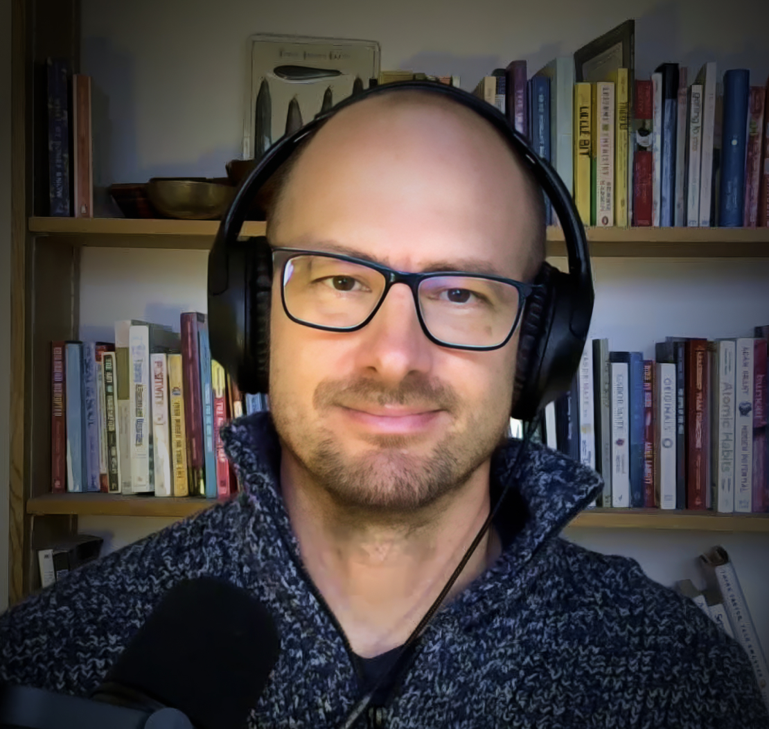 A person wearing headphones and glasses sits in front of a bookshelf, speaking into a microphone.
