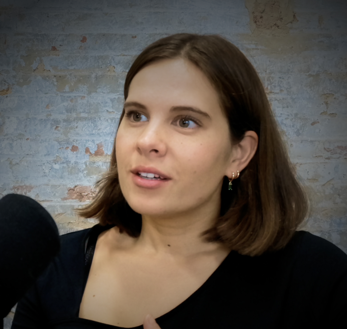 Woman with brown hair speaking into a microphone against a textured wall background.