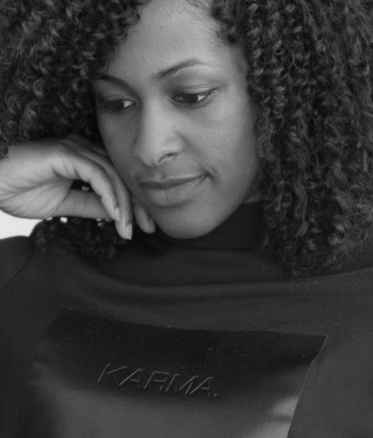 Black and white image of a person with curly hair, looking down thoughtfully, wearing a sweater with "KARMA" written on it.