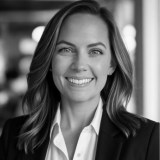 A person with shoulder-length hair smiles, wearing a blazer and a white shirt. The background is softly blurred.