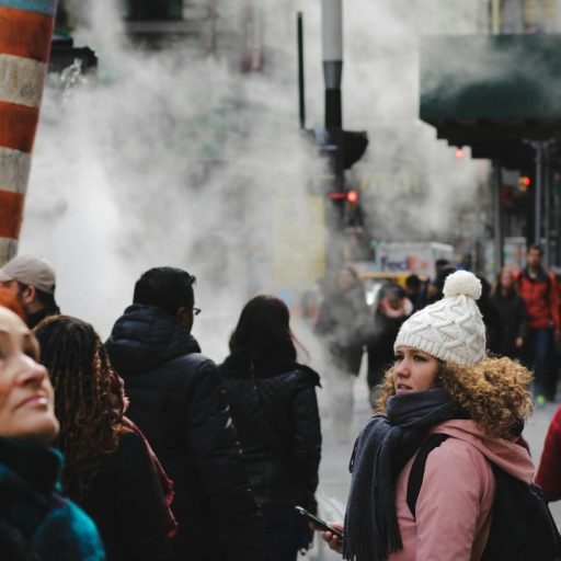 People walking on a city street with steam rising from vents create a scene reminiscent of an omics exposome research study. A woman in a white beanie looks back as buildings and traffic form the vibrant backdrop.