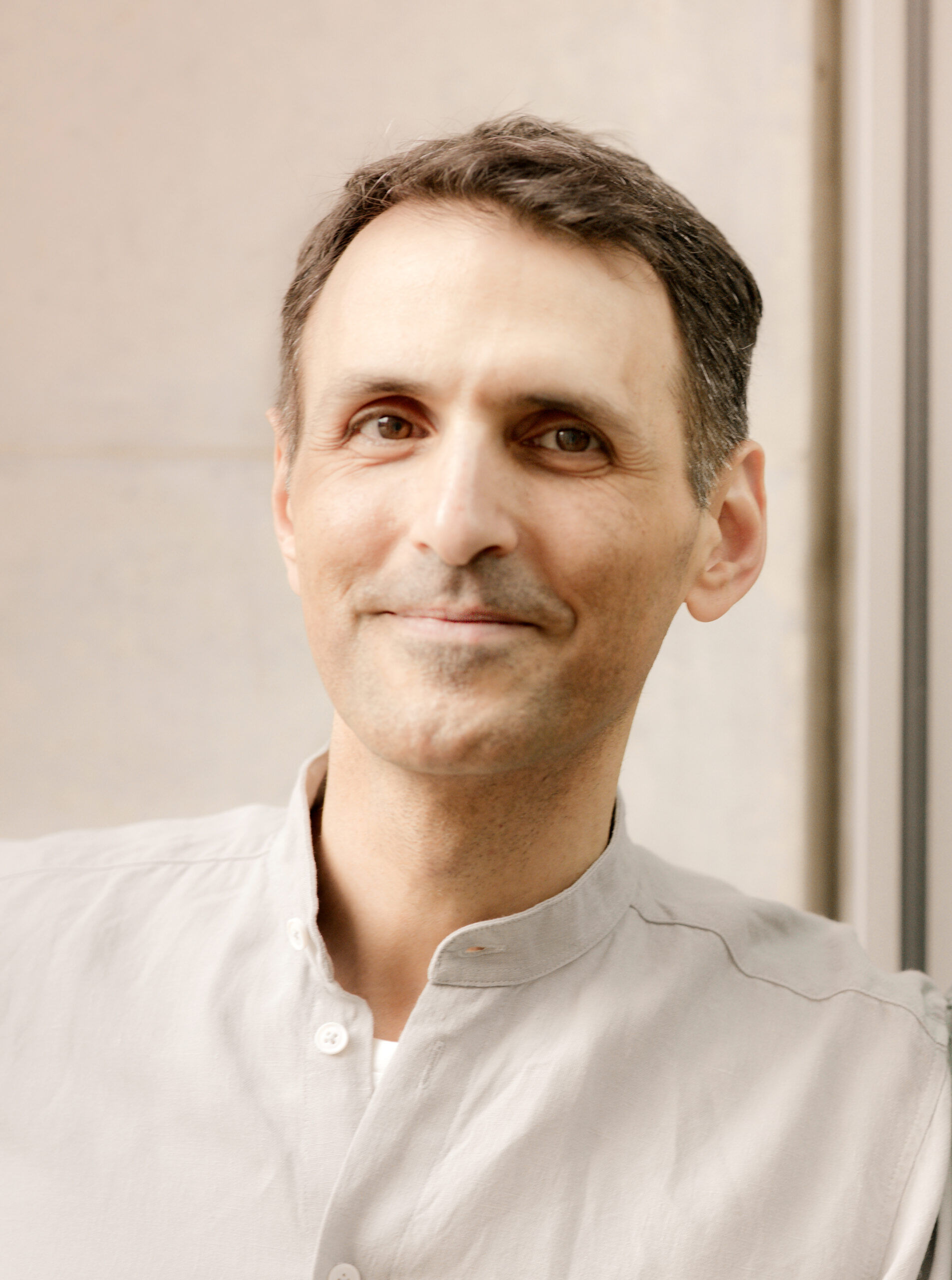 A person with short hair wearing a light-colored shirt is smiling slightly while standing indoors.