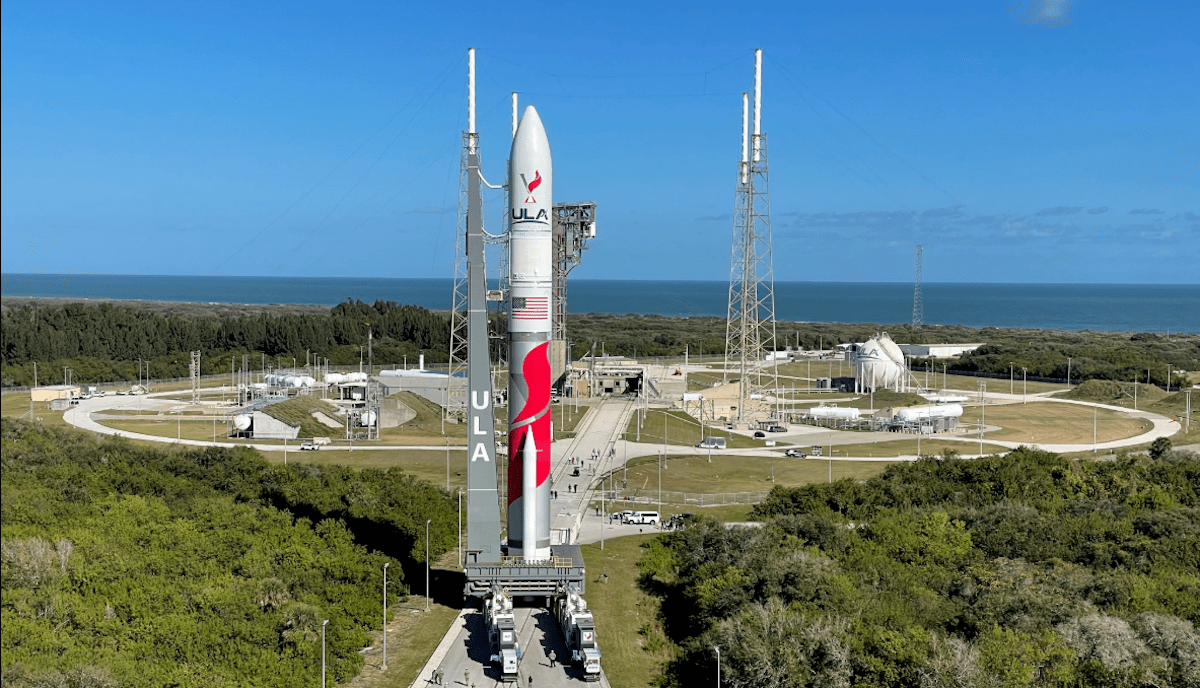 A rocket stands on a launch pad, surrounded by greenery and blue sky, with the ocean in the background.